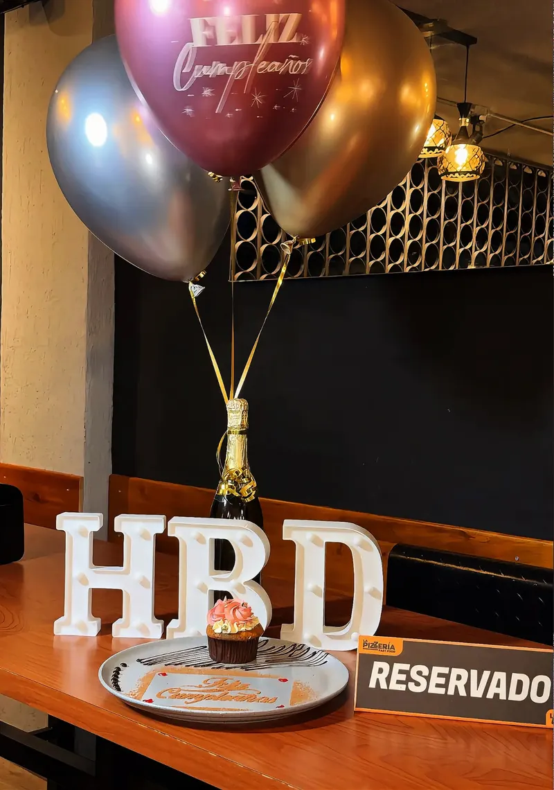 Mesa de cumpleaños con letrero HBD, globos en helio y postre en La Pizzería Fast Food
